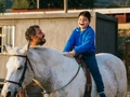 Un enfant aveugle chevauche un cheval avec un grand sourire. À côté, un homme l'accompagne.